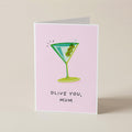A folded greeting card standing upright on a light beige surface. The card has a pale pink background with a cartoon martini glass illustration in green and blue, plus the text 'OLIVE YOU, MUM' in black handwritten-style lettering.