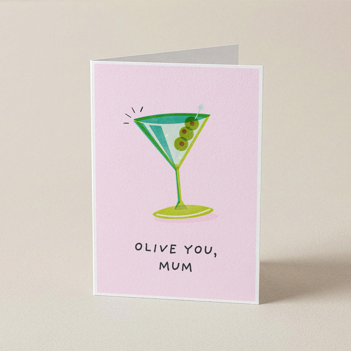 A folded greeting card standing upright on a light beige surface. The card has a pale pink background with a cartoon martini glass illustration in green and blue, plus the text 'OLIVE YOU, MUM' in black handwritten-style lettering.
