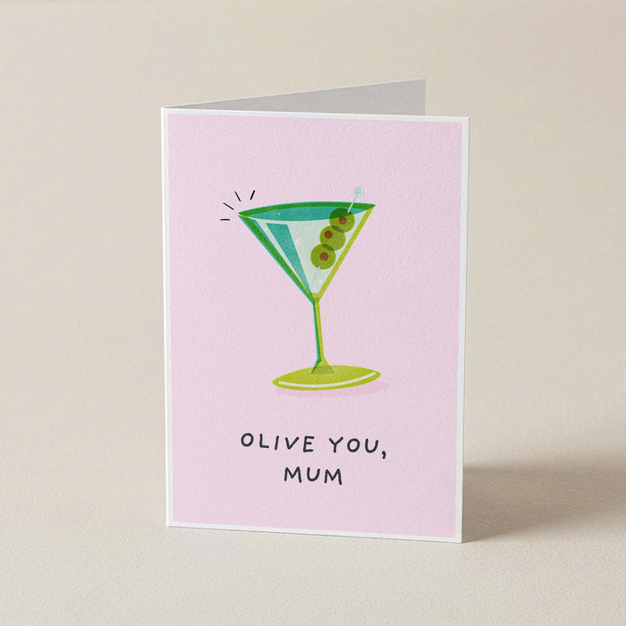A folded greeting card standing upright on a light beige surface. The card has a pale pink background with a cartoon martini glass illustration in green and blue, plus the text 'OLIVE YOU, MUM' in black handwritten-style lettering.