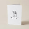 A white folded greeting card standing upright on a light beige surface. The front has a simple black line drawing of a bride and groom holding hands, with the words 'here's to forever!' printed below.