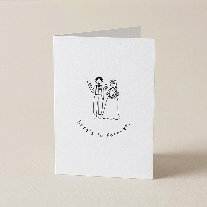 A white folded greeting card standing upright on a light beige surface. The front has a simple black line drawing of a bride and groom holding hands, with the words 'here's to forever!' printed below.