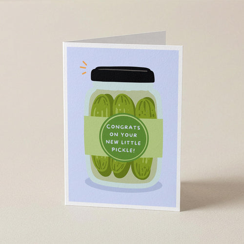 A folded greeting card standing upright on a light beige surface. The card features an illustration of three pickles in a glass jar with a black lid and a green label that reads, 'CONGRATS ON YOUR NEW LITTLE PICKLE!'.