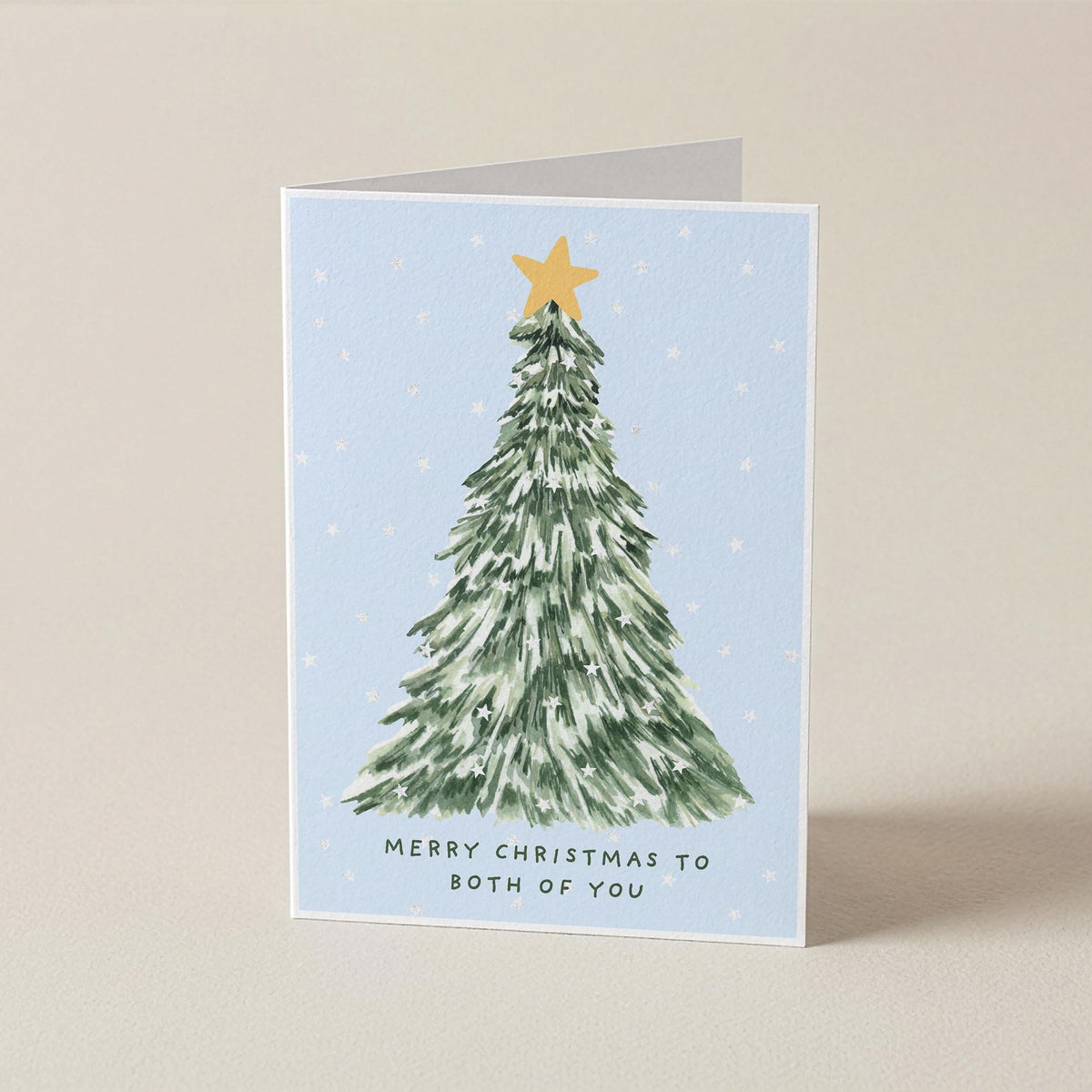 A folded Christmas greeting card standing upright on a light beige surface. The card features a watercolor-style green Christmas tree with a gold star on a pale blue background, with the text 'MERRY CHRISTMAS TO BOTH OF YOU' printed below.