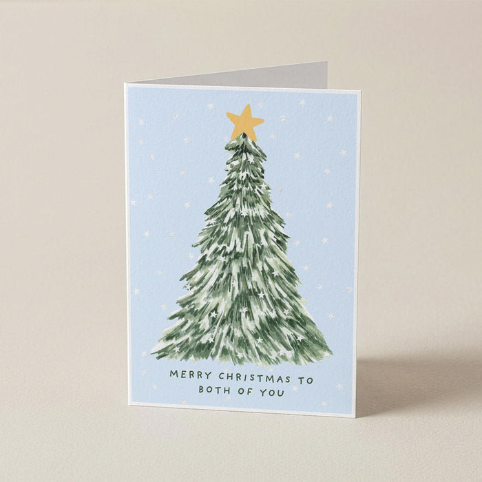 A folded Christmas greeting card standing upright on a light beige surface. The card features a watercolor-style green Christmas tree with a gold star on a pale blue background, with the text 'MERRY CHRISTMAS TO BOTH OF YOU' printed below.