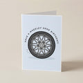 A folded greeting card standing upright on a light beige surface. The card has a white background with a printed illustration of a gray and black wheel in the center and the text 'HAVE A WHEELEY GOOD BIRTHDAY' curved above it.
