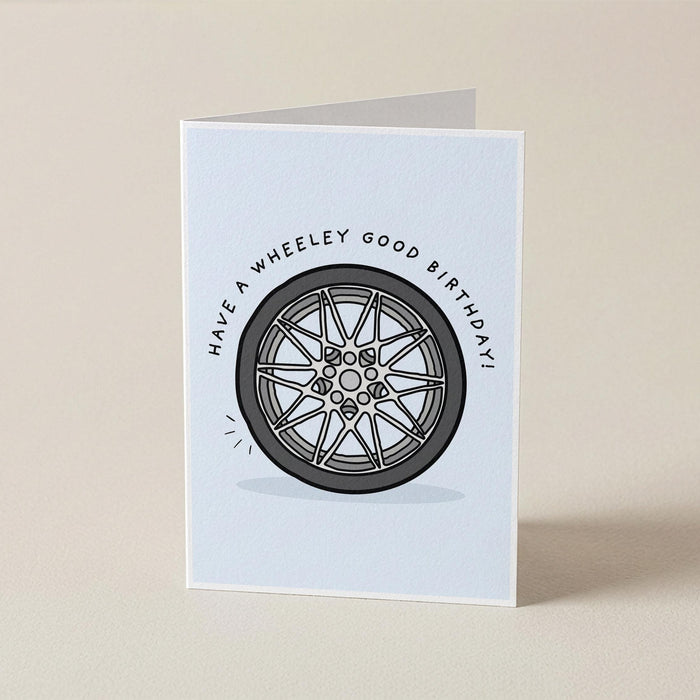 A folded greeting card standing upright on a light beige surface. The card has a white background with a printed illustration of a gray and black wheel in the center and the text 'HAVE A WHEELEY GOOD BIRTHDAY' curved above it.