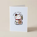 A folded greeting card standing upright on a light beige surface. The front of the card features a white background with a cute illustrated maneki-neko cat in black, white, red, and gold, with the words 'GOOD LUCK!' arched above it.