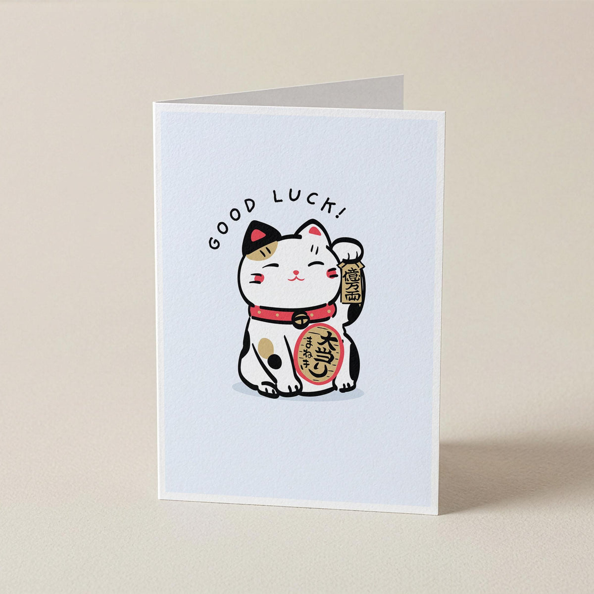A folded greeting card standing upright on a light beige surface. The front of the card features a white background with a cute illustrated maneki-neko cat in black, white, red, and gold, with the words 'GOOD LUCK!' arched above it.