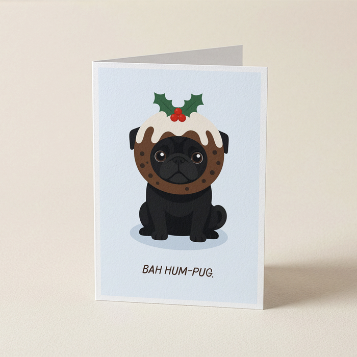 A folded greeting card standing upright on a light beige surface. The card features a cartoon black pug wearing a Christmas pudding decoration with holly on a pale blue background, with the text 'BAH HUM-PUG.