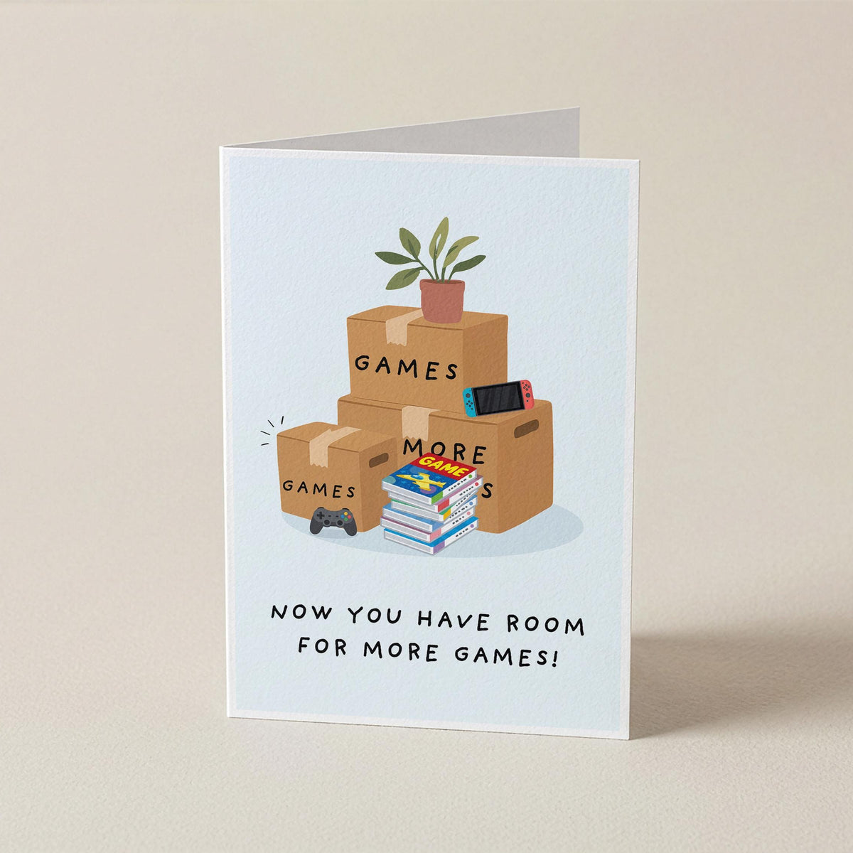 A folded greeting card standing upright on a beige surface. The card features an illustration of stacked cardboard boxes labeled 'GAMES' and 'MORE', with a small potted plant and game controller, above the text 'NOW YOU HAVE ROOM FOR MORE GAMES!'.