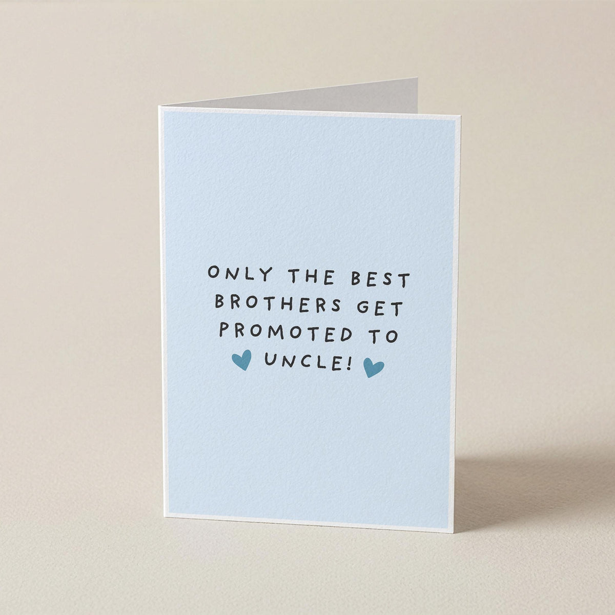 A folded greeting card standing upright on a light beige surface. The card is pale blue with the printed message 'ONLY THE BEST BROTHERS GET PROMOTED TO UNCLE!' in black text and two small blue hearts.