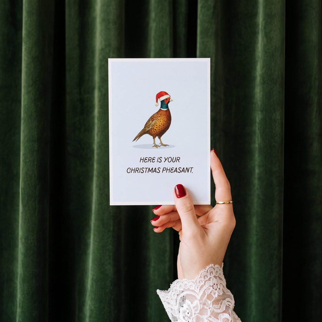 A hand with red nail polish and a white lace cuff holds up a white greeting card featuring an illustration of a pheasant wearing a Santa hat, with the text 'HERE IS YOUR CHRISTMAS PHEASANT.' The background is a dark green velvet curtain.