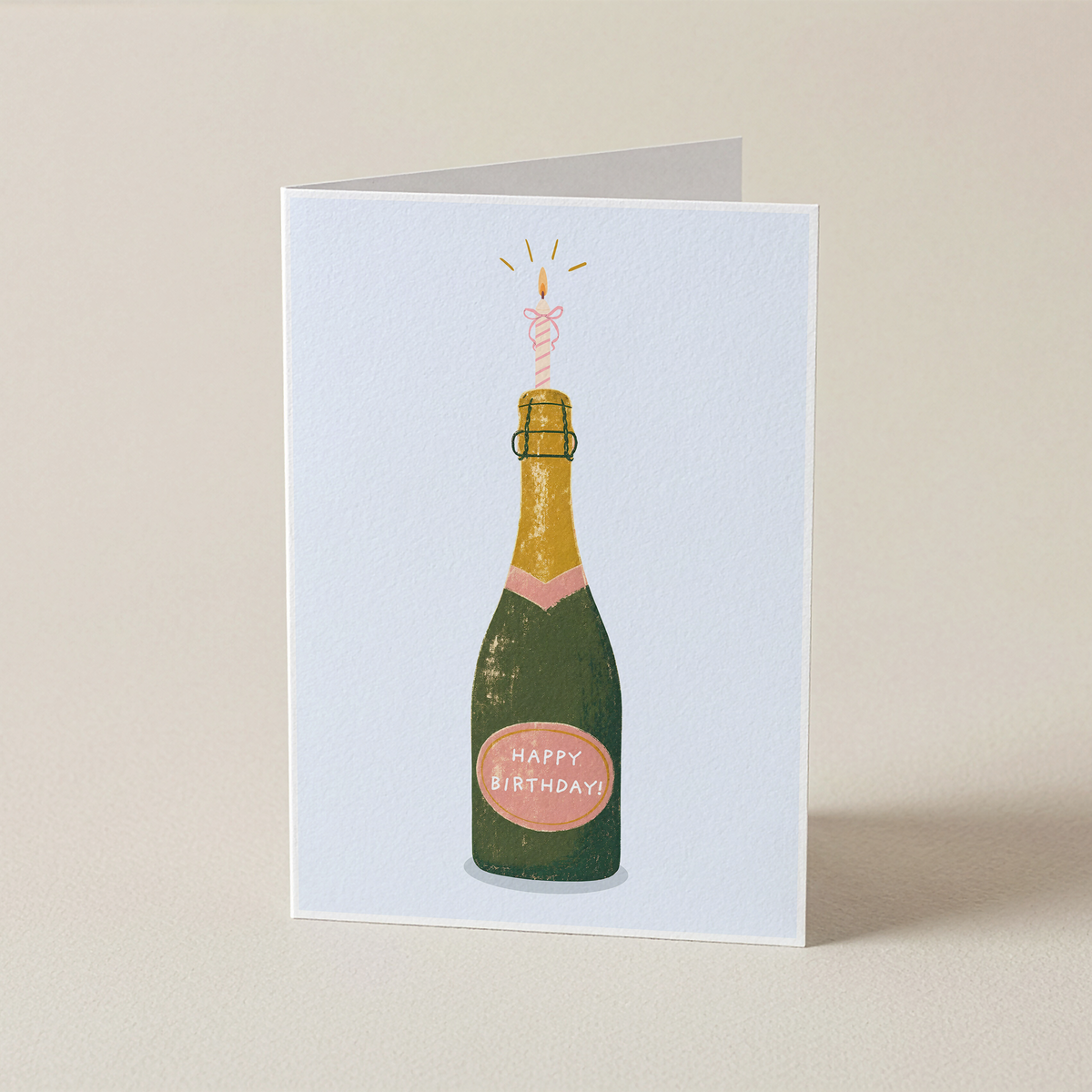 A folded greeting card standing upright on a light beige surface. The card features an illustration of a champagne bottle with a pink sparkler and a pink 'Happy Birthday' label.