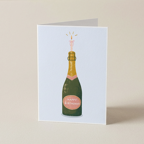 A folded greeting card standing upright on a light beige surface. The card features an illustration of a champagne bottle with a pink sparkler and a pink 'Happy Birthday' label.