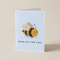 A folded greeting card standing upright on a light beige background. The card has a pale blue front featuring a cute cartoon bee and the text 'BUZZING FOR YOU!'.
