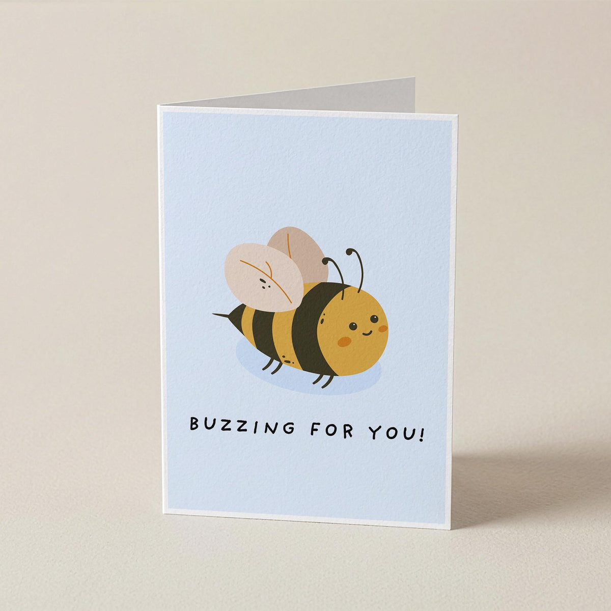 A folded greeting card standing upright on a light beige background. The card has a pale blue front featuring a cute cartoon bee and the text 'BUZZING FOR YOU!'.