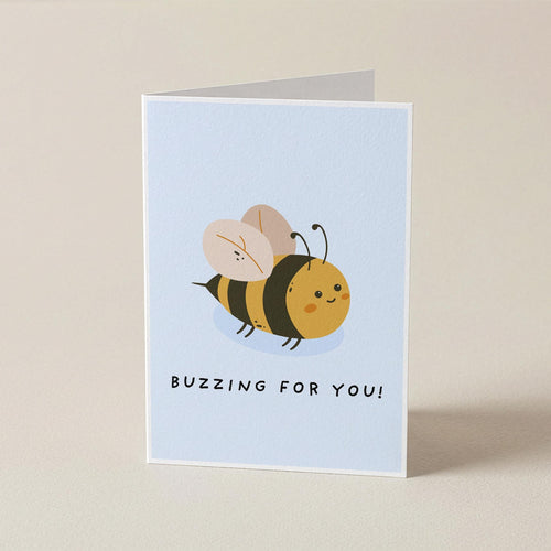 A folded greeting card standing upright on a light beige background. The card has a pale blue front featuring a cute cartoon bee and the text 'BUZZING FOR YOU!'.