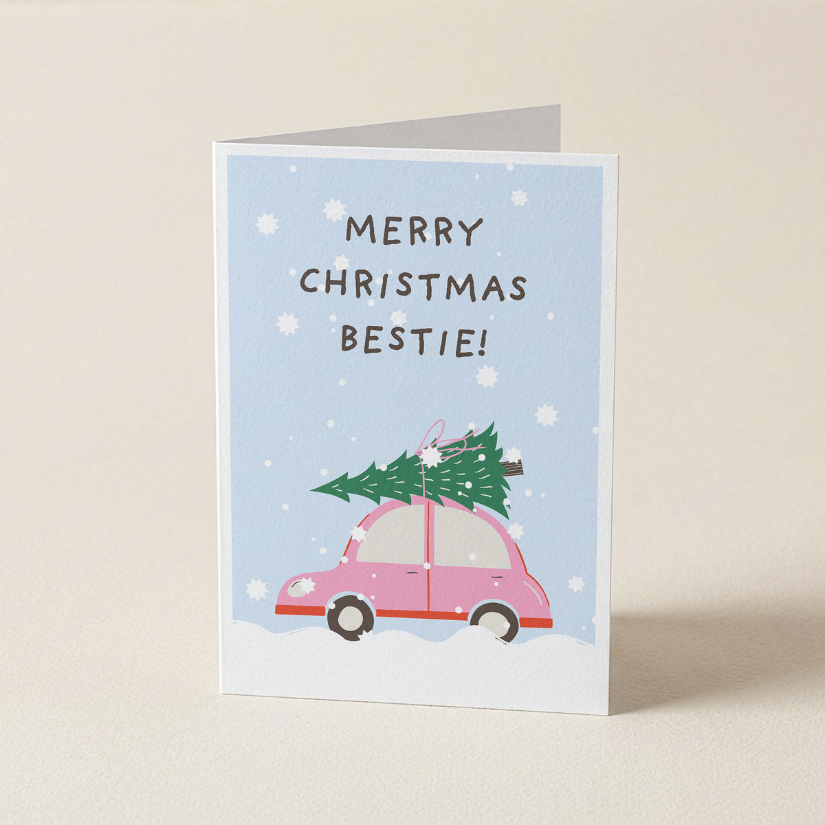 A folded Christmas greeting card standing upright on a light beige surface. The card shows a light blue winter scene with the text 'MERRY CHRISTMAS BESTIE!' above a pink car carrying a green Christmas tree.