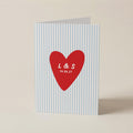 A personalized greeting card with a light blue and white vertical stripe pattern, featuring a red heart in the center with the white printed initials 'L & S' and the date '19.06.