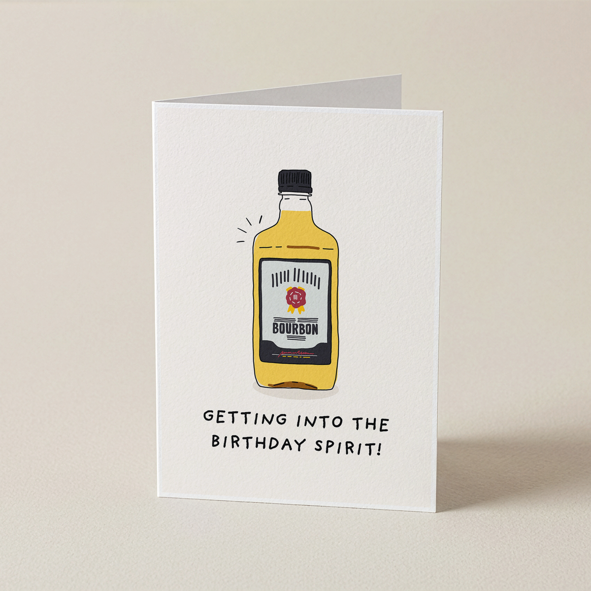 A folded white greeting card standing upright on a light beige surface. The card features a hand-drawn illustration of a yellow bourbon bottle with the words 'GETTING INTO THE BIRTHDAY SPIRIT!' printed below in black.
