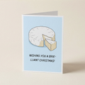 A folded greeting card standing upright on a light beige surface. The card has a pale blue center with an illustration of a sliced brie cheese and the text 'WISHING YOU A BRIE-LLIANT CHRISTMAS!'.