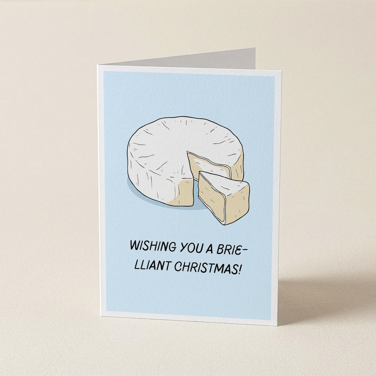 A folded greeting card standing upright on a light beige surface. The card has a pale blue center with an illustration of a sliced brie cheese and the text 'WISHING YOU A BRIE-LLIANT CHRISTMAS!'.