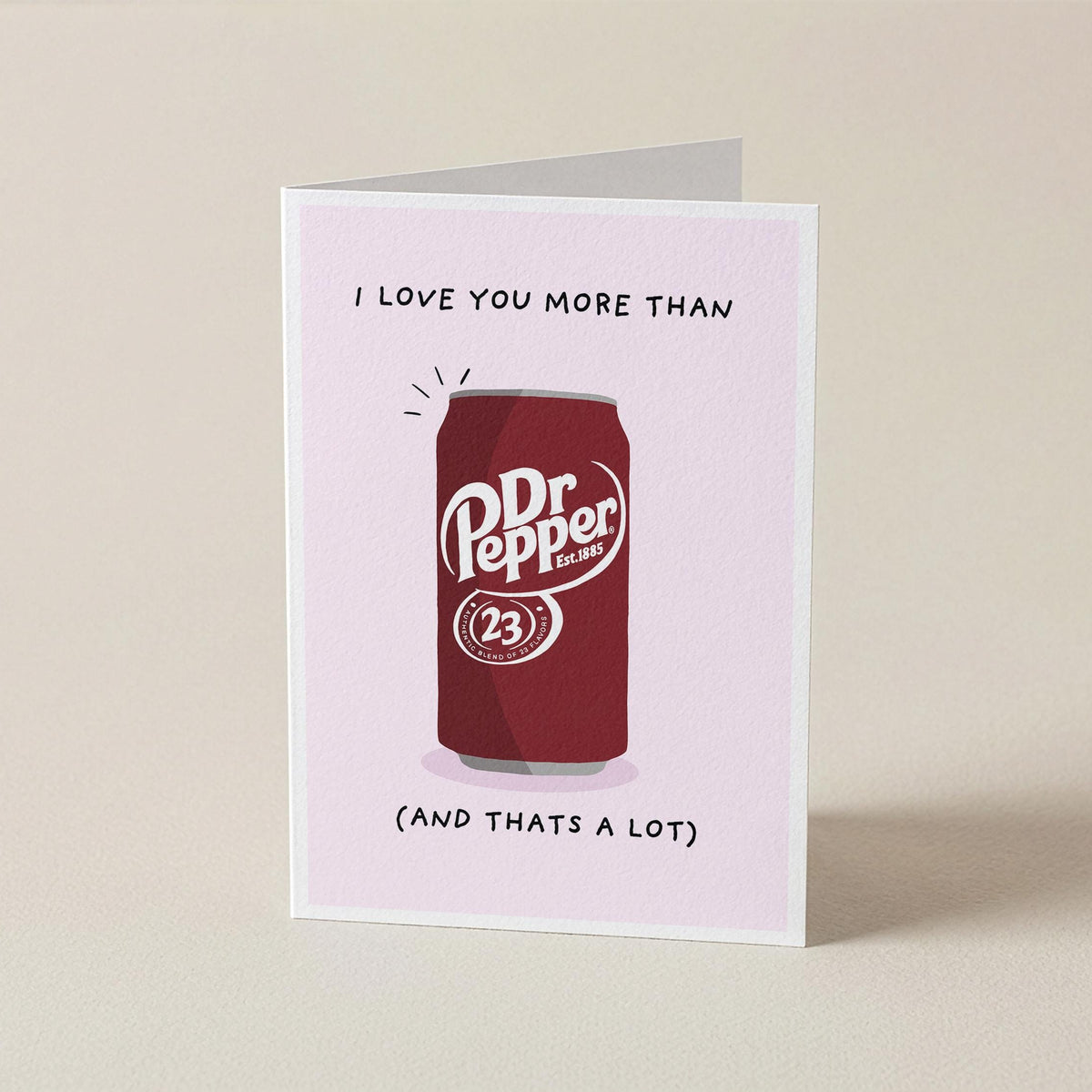 A folded greeting card standing upright on a light beige surface. The card has a pale pink background and features a red Dr. Pepper can.