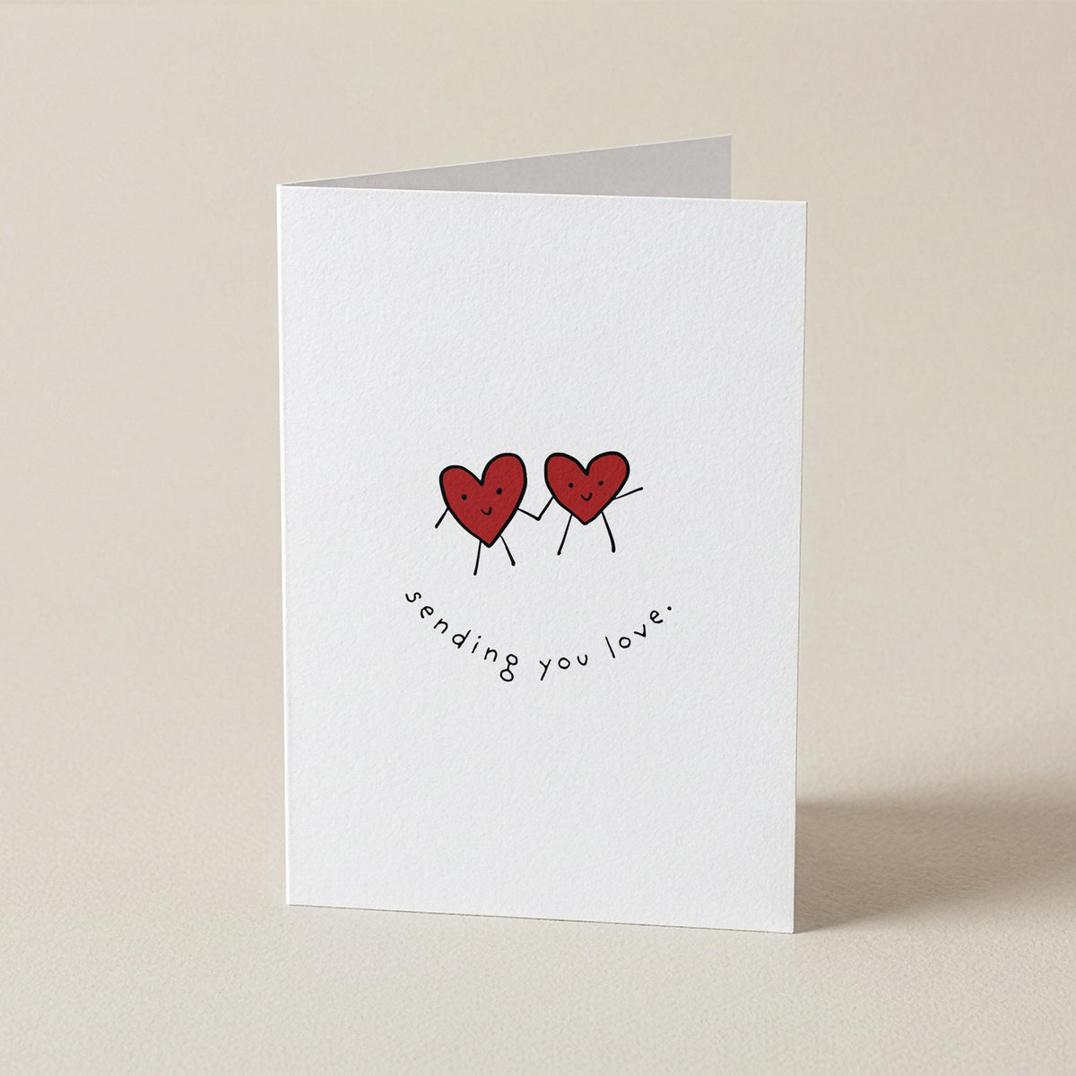 A white greeting card standing upright on a light beige surface, featuring two small red hearts with thin black limbs and the handwritten text 'sending you love.' below them.