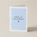 A square greeting card with a white border and a light blue scalloped center, reading 'HAPPY 1ST ANNIVERSARY' above a small red heart, photographed on a plain beige background.