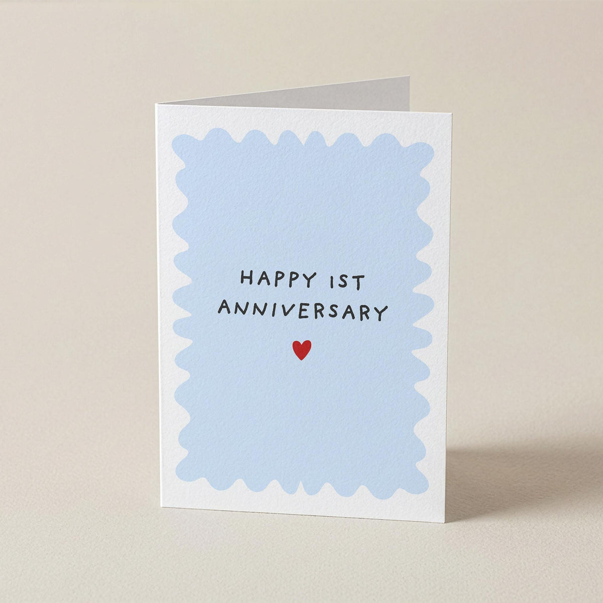 A square greeting card with a white border and a light blue scalloped center, reading 'HAPPY 1ST ANNIVERSARY' above a small red heart, photographed on a plain beige background.