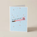 A folded greeting card standing upright on a light beige surface. The card has a pale blue background with scattered pink, white, and brown confetti, a pink party blower illustration with the word 'woohoo!' across it, and a white border.