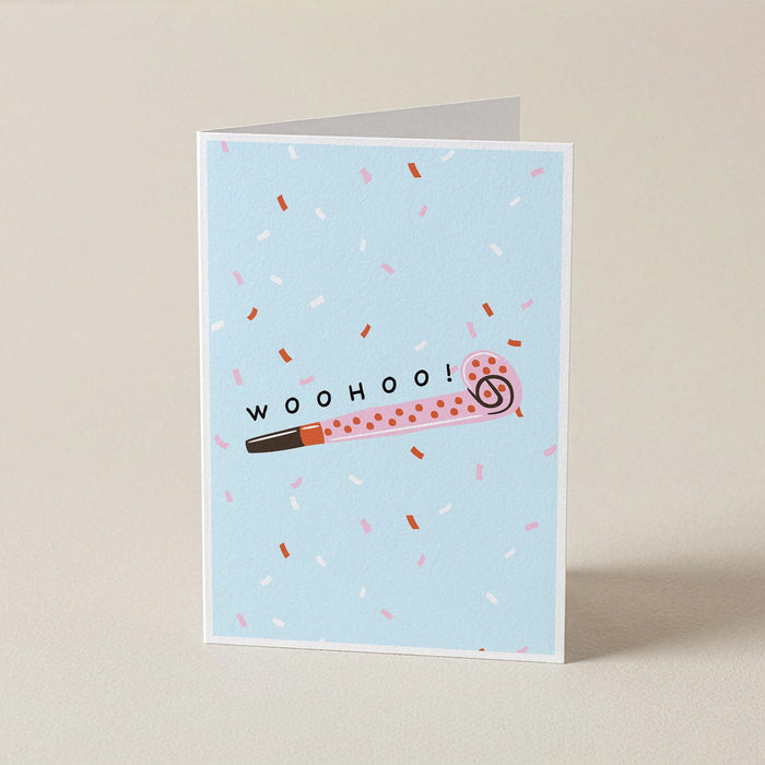 A folded greeting card standing upright on a light beige surface. The card has a pale blue background with scattered pink, white, and brown confetti, a pink party blower illustration with the word 'woohoo!' across it, and a white border.