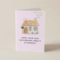 A folded greeting card standing upright on a light beige surface. The card has a pale pink background with a small illustrated house in the center and the text 'HOPE YOUR NEW NEIGHBORS AREN'T DICKHEADS' printed below.