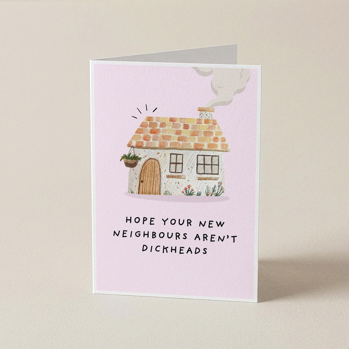 A folded greeting card standing upright on a light beige surface. The card has a pale pink background with a small illustrated house in the center and the text 'HOPE YOUR NEW NEIGHBORS AREN'T DICKHEADS' printed below.