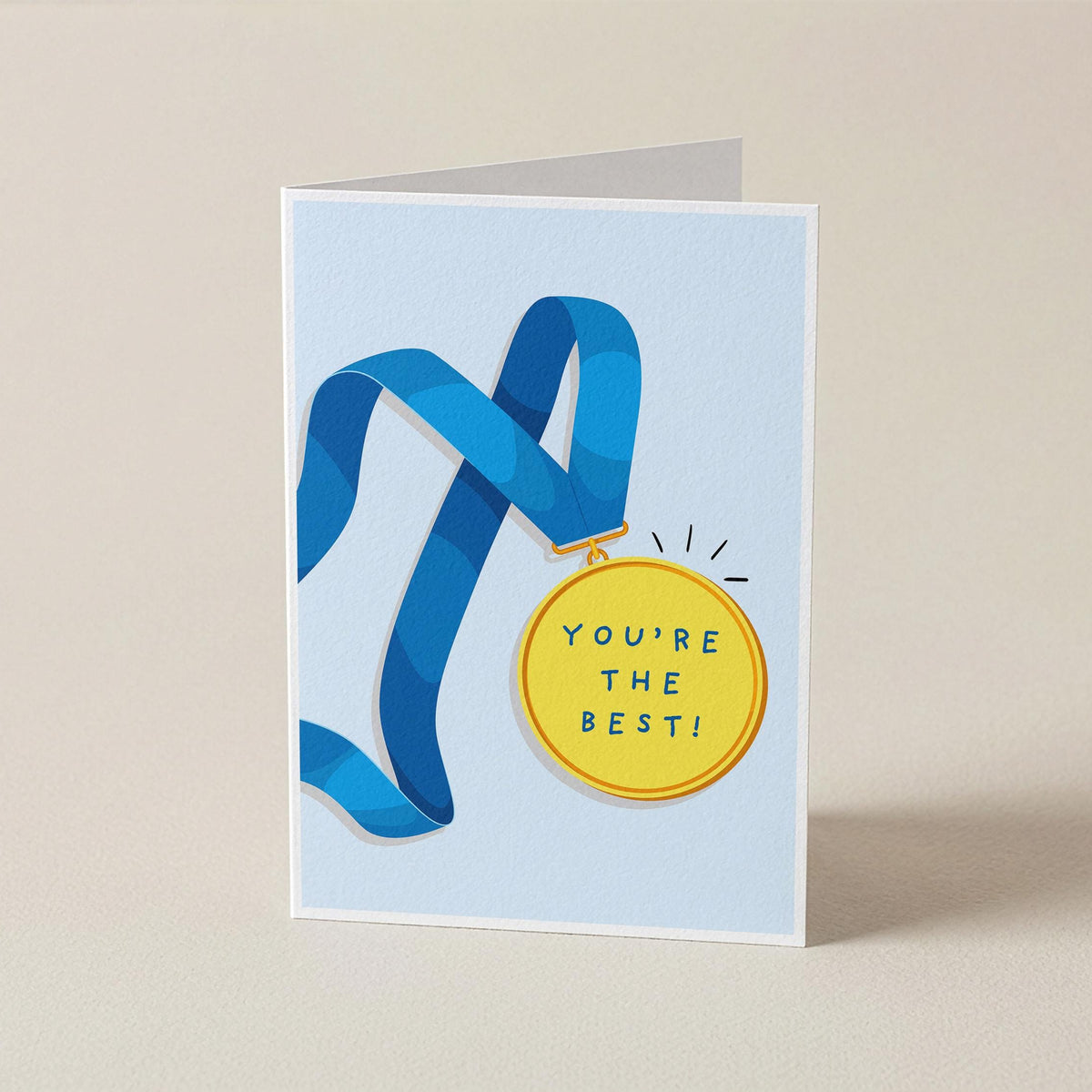 A folded greeting card standing upright on a light beige surface. The card has a pale blue background with a blue ribbon and a yellow medallion graphic, plus the text 'YOU'RE THE BEST!' on the medallion.