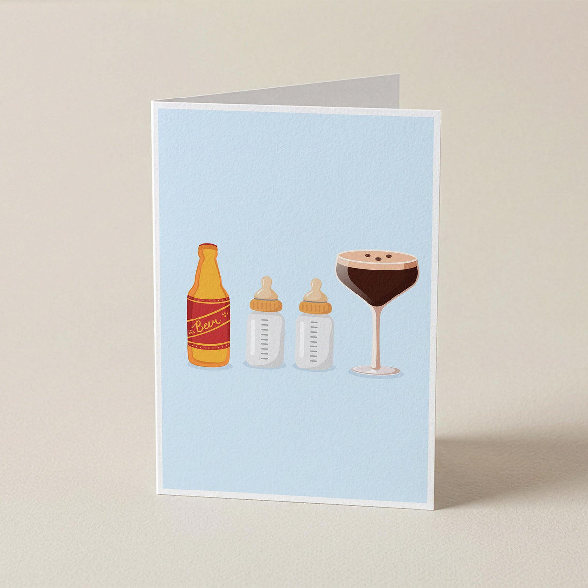 A folded greeting card standing upright on a beige surface, featuring a light blue background with illustrated baby bottle and cocktail glass graphics.