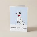 A folded Christmas greeting card standing upright on a light beige surface. The card features an illustration of a Dalmatian dog in a snowy scene against a pale blue background, with the text 'MERRY CHRISTMAS!' printed at the bottom.