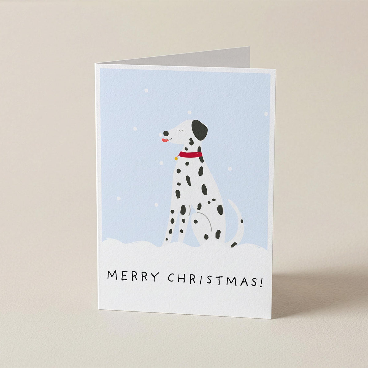 A folded Christmas greeting card standing upright on a light beige surface. The card features an illustration of a Dalmatian dog in a snowy scene against a pale blue background, with the text 'MERRY CHRISTMAS!' printed at the bottom.