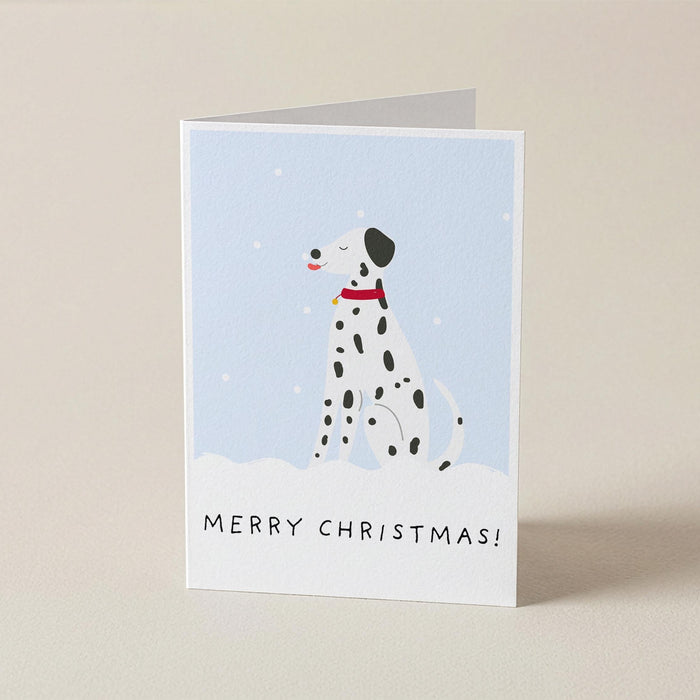 A folded Christmas greeting card standing upright on a light beige surface. The card features an illustration of a Dalmatian dog in a snowy scene against a pale blue background, with the text 'MERRY CHRISTMAS!' printed at the bottom.