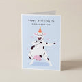 A folded greeting card standing upright on a light beige surface. The card has a pale blue front with the text 'happy birthday to moooooooooo' above a cartoon cow dancing and holding a pink ball.