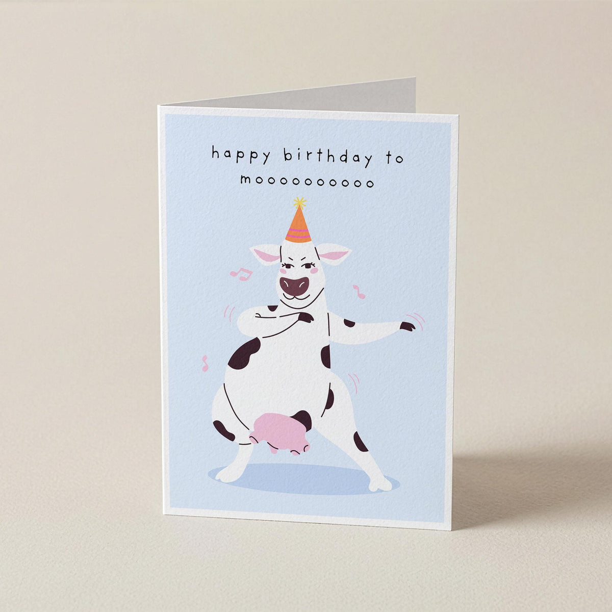 A folded greeting card standing upright on a light beige surface. The card has a pale blue front with the text 'happy birthday to moooooooooo' above a cartoon cow dancing and holding a pink ball.
