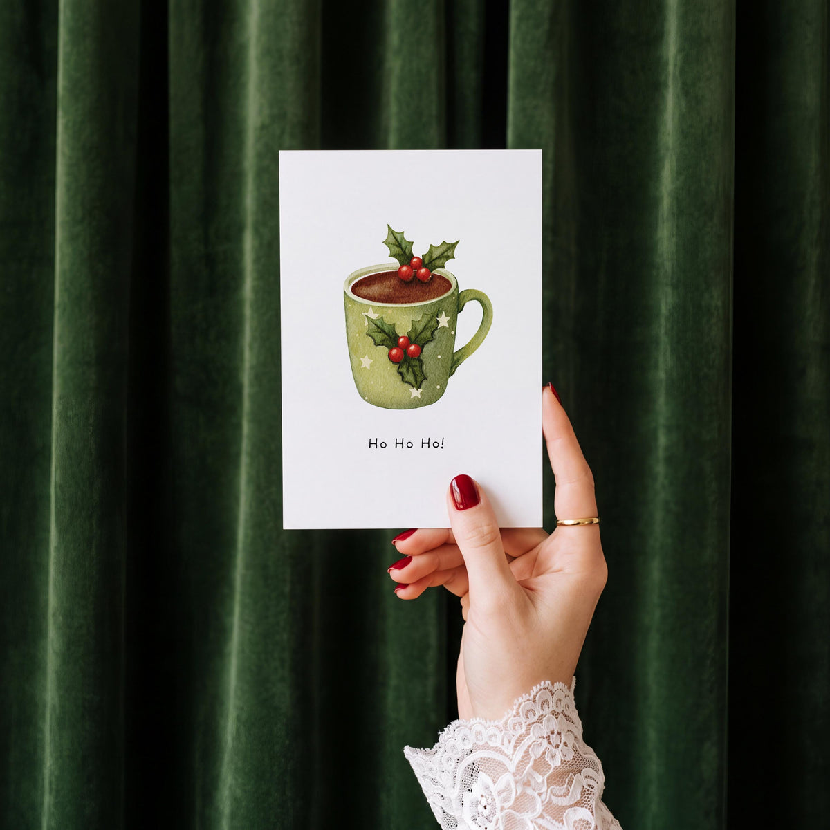 A hand with red nail polish and a white lace cuff holds up a white greeting card featuring an illustration of a green mug filled with hot cocoa, decorated with holly leaves and red berries, with the text 'Ho Ho Ho!' underneath. The background is a dark green velvet curtain.