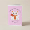 A folded greeting card standing upright on a light beige surface. The card has a pink background with a cartoon white cat holding an orange fan and wearing an orange collar, with the text 'WISHING YOU ALL THE LUCK IN THE WORLD!'.