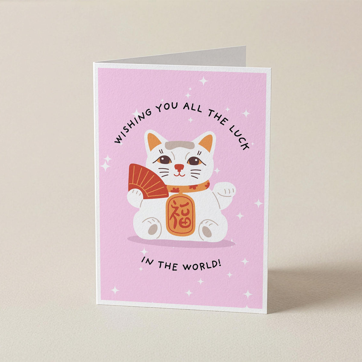 A folded greeting card standing upright on a light beige surface. The card has a pink background with a cartoon white cat holding an orange fan and wearing an orange collar, with the text 'WISHING YOU ALL THE LUCK IN THE WORLD!'.