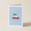 A folded greeting card standing upright on a light beige surface. The card has a white border and a light blue scalloped center featuring a red car carrying a green Christmas tree, with white snowflakes scattered throughout.