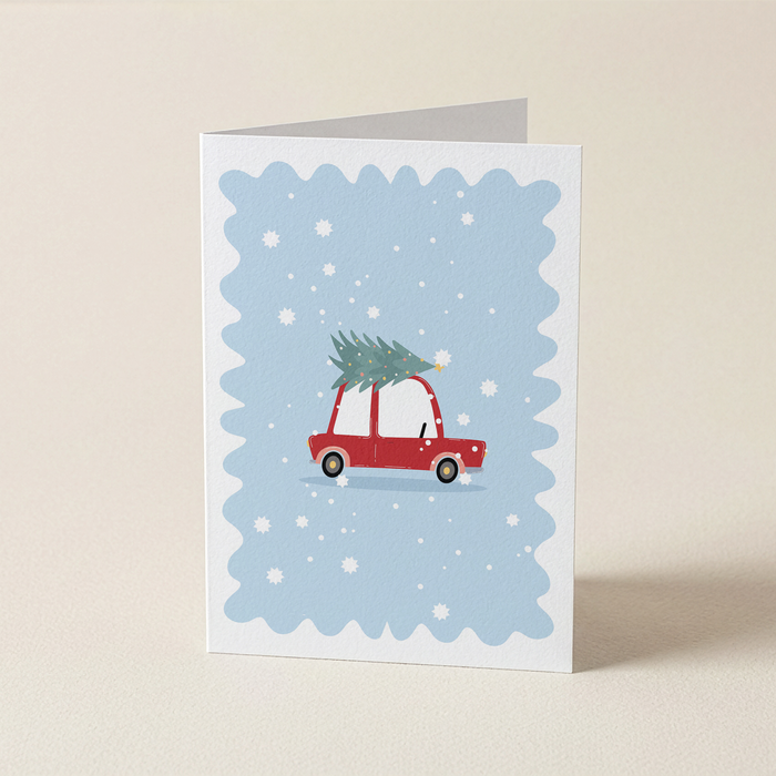 A folded greeting card standing upright on a light beige surface. The card has a white border and a light blue scalloped center featuring a red car carrying a green Christmas tree, with white snowflakes scattered throughout.