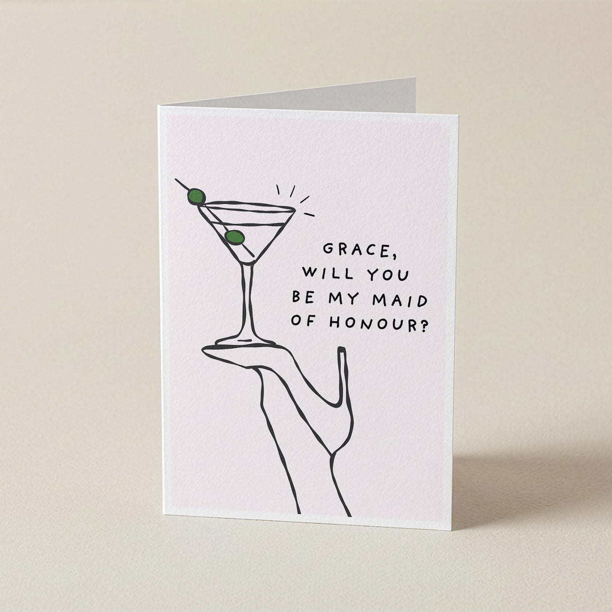 A white greeting card with a minimalist black line drawing of a hand holding a martini glass with two green olives, next to the text 'GRACE, WILL YOU BE MY MAID OF HONOUR?'. The card is standing upright on a light beige surface against a matching beige background.