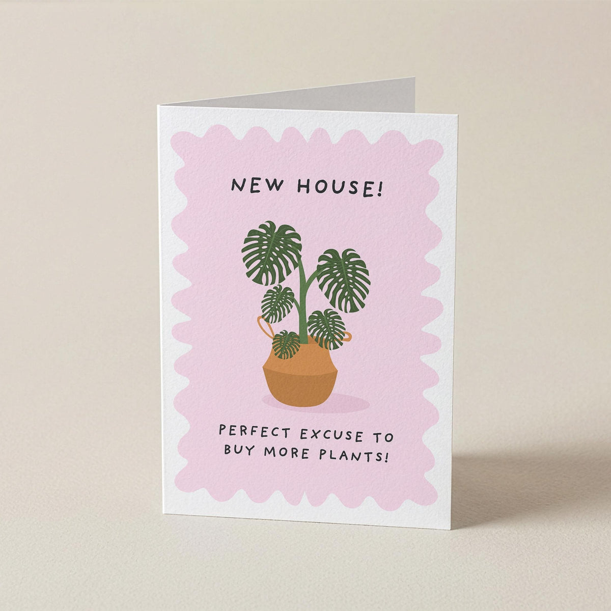 A folded greeting card standing upright on a light beige surface. The card has a white border with a pink scalloped center, featuring an illustration of potted green plants and the text 'NEW HOUSE! PERFECT EXCUSE TO BUY MORE PLANTS!'.