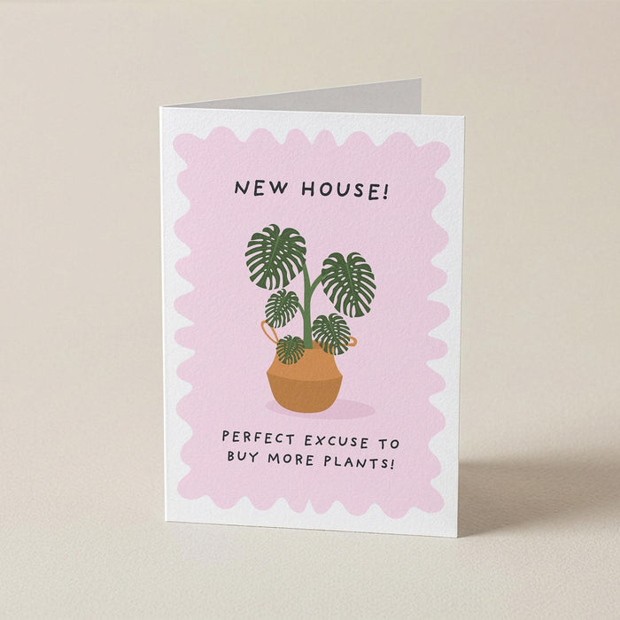 A folded greeting card standing upright on a light beige surface. The card has a white border with a pink scalloped center, featuring an illustration of potted green plants and the text 'NEW HOUSE! PERFECT EXCUSE TO BUY MORE PLANTS!'.