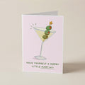 A folded greeting card standing upright on a light beige surface. The card has a pale pink background and features an illustration of a martini glass with green olives and a small gold star, with the text 'HAVE YOURSELF A MERRY LITTLE MARTINI!!' printed below.